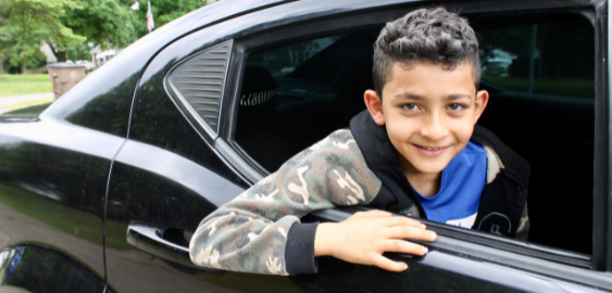Image of a grinning boy with dark hair leaning his head outside of a stopped black car.